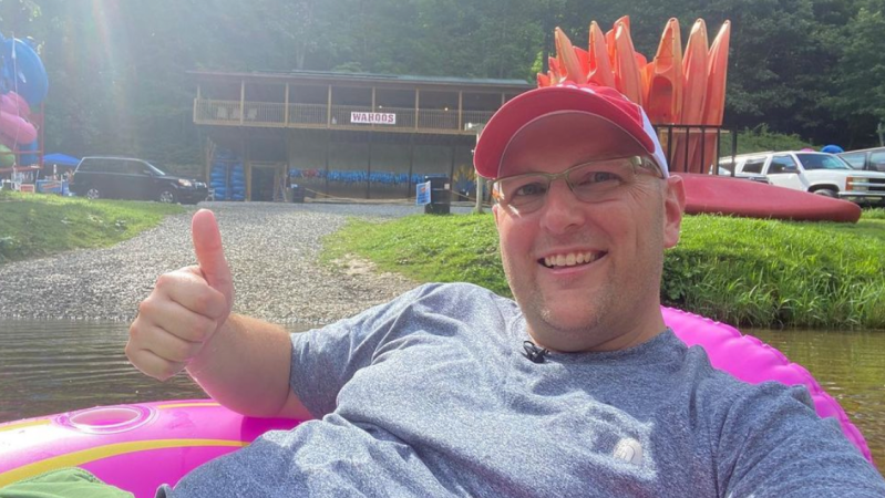 Smiling man in a cap gives a thumbs up while river tubing in a pink tube near a rental shop.