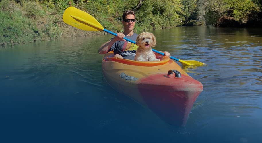 Man paddling a kayak with his dog, passing treehouse cabins along a calm, scenic river.