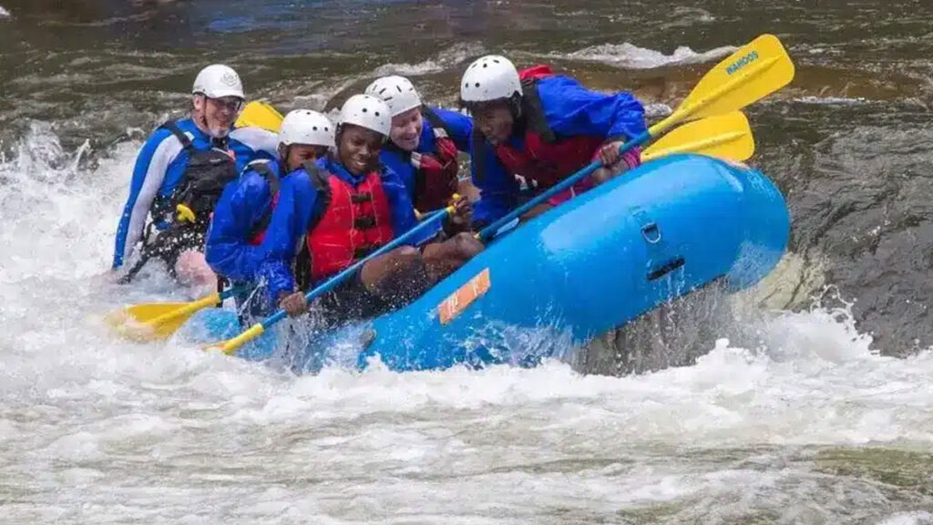 Six people river tubing in a blue raft, wearing helmets and life jackets on a wild river.
