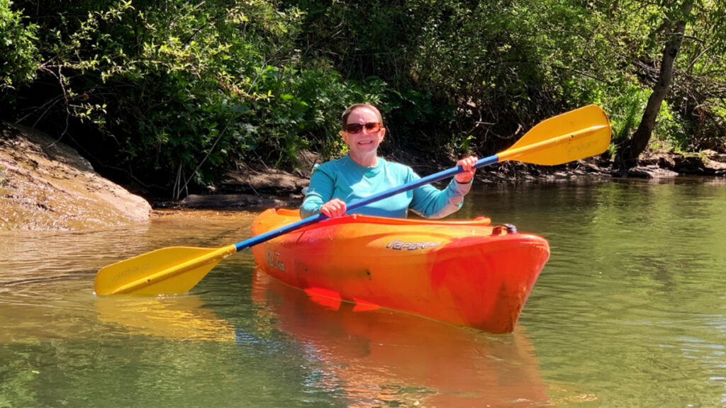 Person kayaking on calm, clear water near a leafy shoreline, perfect for camping adventures.