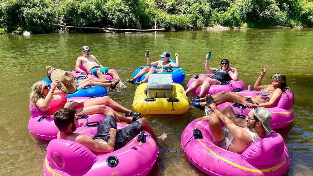 Six people enjoy river tubing, relaxing in colorful tubes, drinks in hand, on a sunny day.