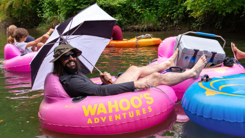 Man lounging on a pink tube with an umbrella, river tubing with others floating nearby.