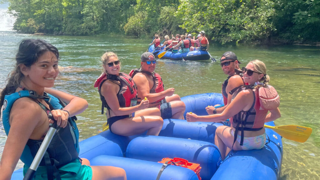 A group of people smiling while river rafting on a scenic river with trees in the background.