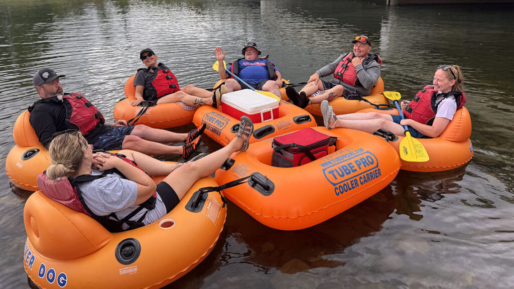 Six people relax in orange tubes on calm water, linked around a cooler after river rafting.