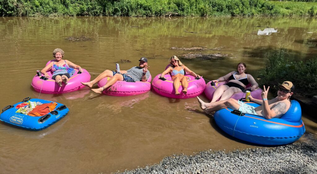 Five adults relax in colorful inner tubes on a river, smiling and enjoying a sunny day.