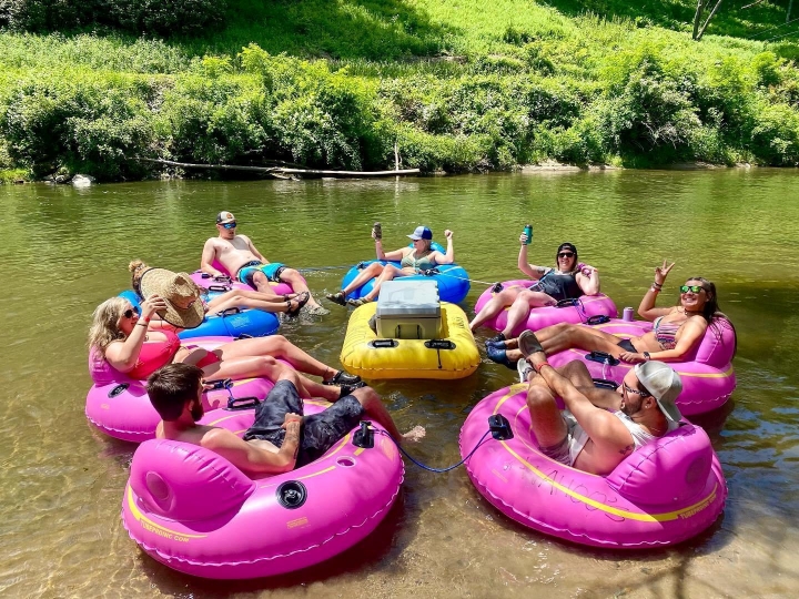 Seven people relax on pink and yellow inner tubes, floating together on a river in the sun.