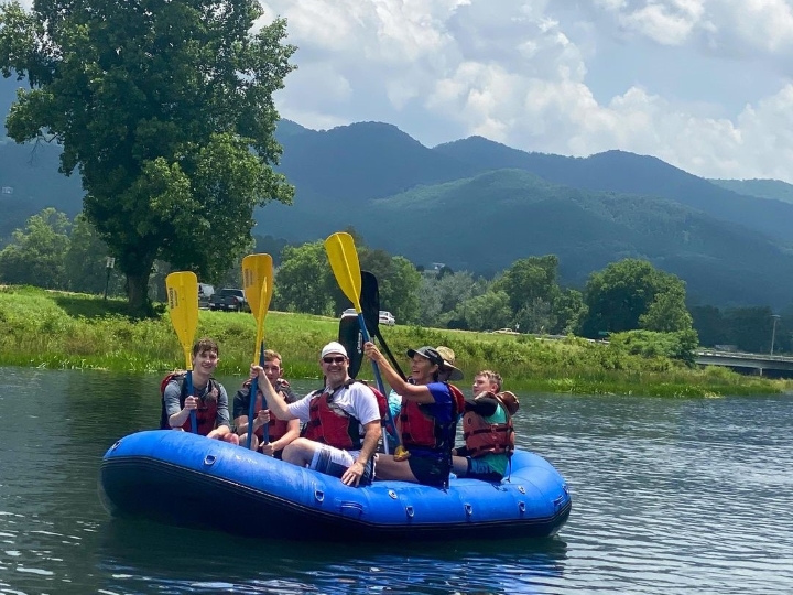 Six people in a blue raft paddle on a calm river with mountains and trees in the background.