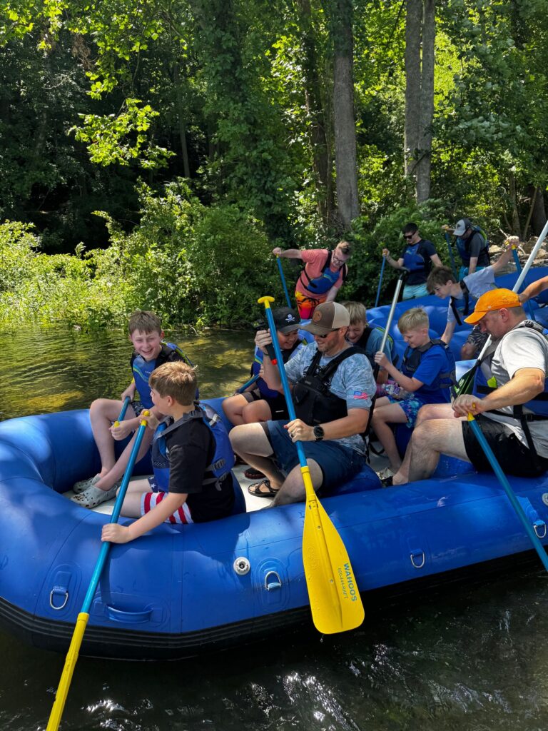 Group of kids and adults in life vests paddling a blue raft on a river, surrounded by trees.
