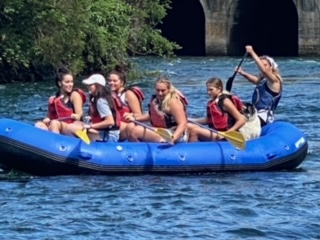 Six people in life jackets paddle a blue inflatable raft on a river near a bridge and greenery.