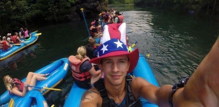 Man in a red, white, and blue hat takes a selfie while rafting with a group on a river.