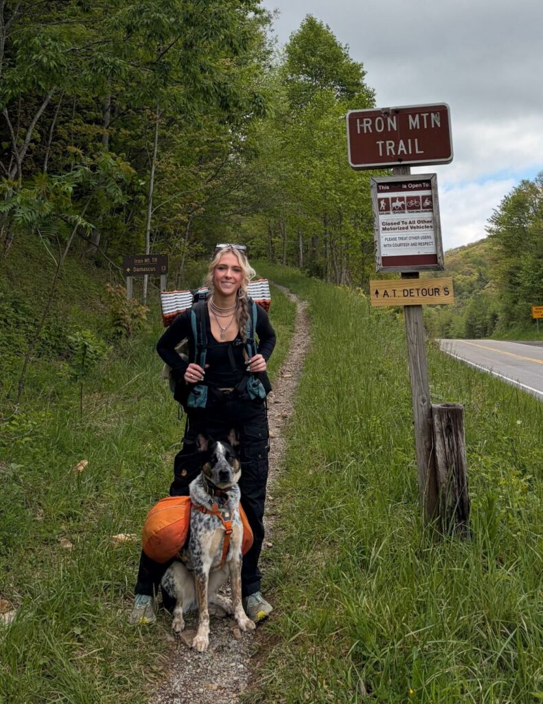 A hiker with a backpack and dog stands by the Iron Mountain Trail sign on a grassy path.