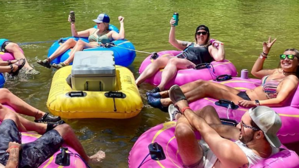 Five people relax on colorful tubes in a river, holding drinks and smiling on a sunny day.