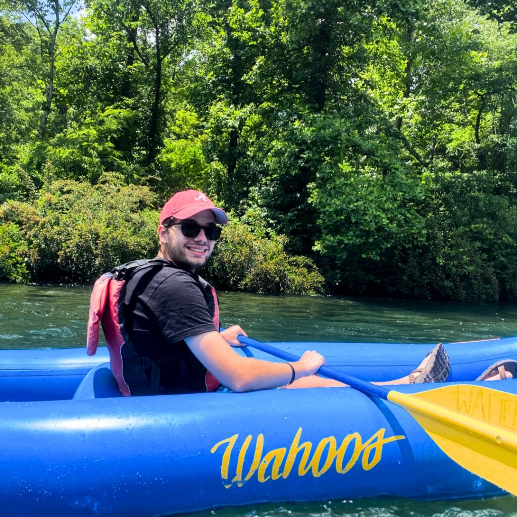 Person kayaking on a blue Wahoos kayak in sunny, green, tree-lined water.