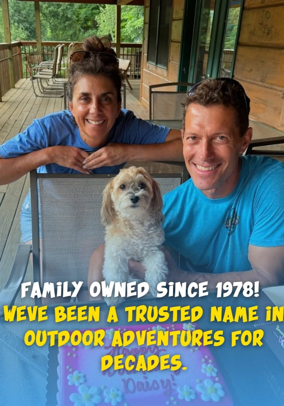 Two authors and a small dog sit outdoors by a table with a pink cake that says Daisy.